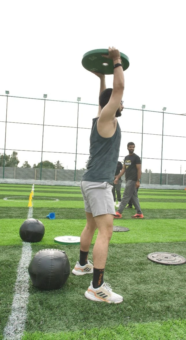 Member pressing a plate overhead beside wall balls on the turf
